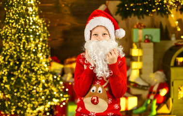 Fluffy beard. Child with white long fake beard near christmas tree. Merry christmas. Happy childhood concept. Kid wear santa hat and christmas sweater. Santa brought me gifts. December tradition