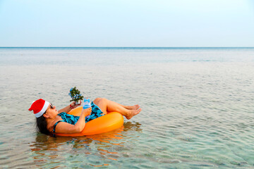 A woman in a swimming ring in a Santa Claus hat with a cocktail and a small Christmas tree in her hand near the shore in the sea. Shooting from the back.