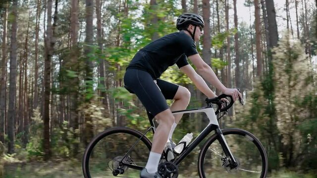 A Male Cyclist Trains By Riding A Road Bike In A Sunny Pine Forest, Wearing Helmet. Workout On Triathlon Bike. Side Shot Of Cyclist In Black Outfit Riding Road Bicycle. 50 Fps. 4k Footage