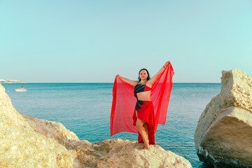 A brunette woman in a red oriental dance costume is dancing on a rock against the backdrop of the sea with a shawl.