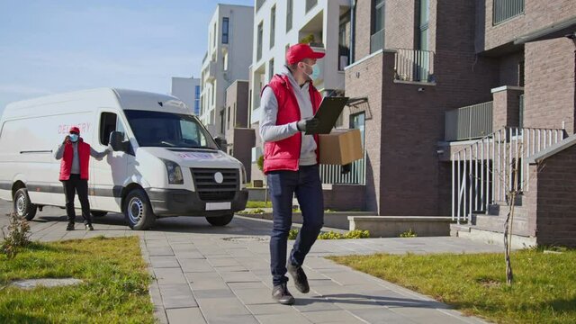 Caucasian delivery man in face mask and gloves handling parcel box, walking along the street with tablet folder. Courier on the way to deliver postal parcel to a client.