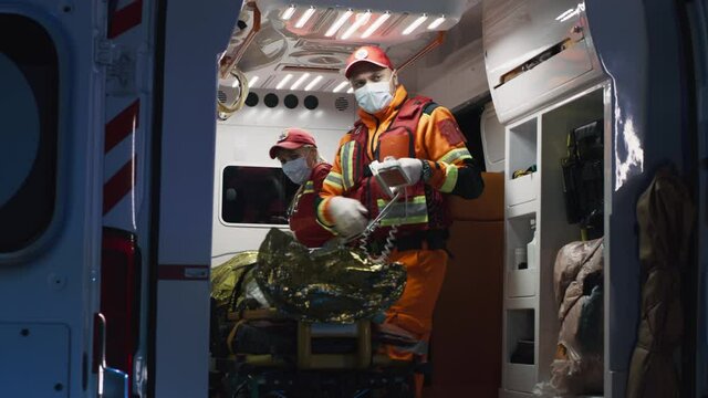 Low angle of men in paramedic uniform preparing defibrillator for patient in emergency vehicle during rescue mission in evening