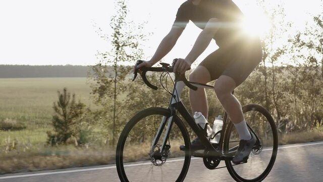 Man riding a road bike at the sunset on a highway near field, wearing helmet. Side shot of cyclist in black outfit riding road bicycle. 50 fps. 4k footage