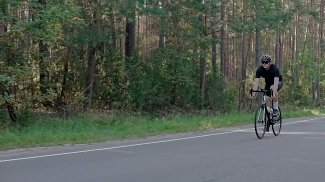 A male cyclist riding a road bike in a sunny pine forest, wearing helmet. Workout on triathlon bike. Side shot of cyclist in black outfit riding road bicycle. 50 fps. 4k footage