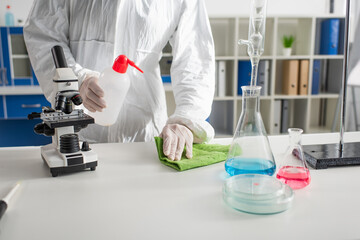 cropped view of doctor in protective suit cleaning desk near microscope in laboratory.