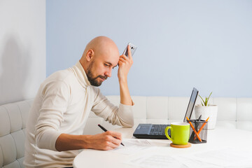 Thouthful bald man with beard in white turtleneck working on his notebook at home, side view