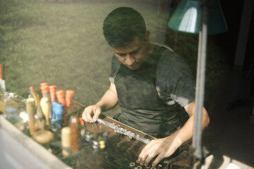 Latin man repairing a flute on his workbench as seen through a window pane