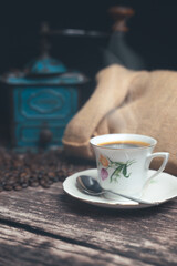 Vertical studio shot of still life with vintage manual green metal coffee grinder, jute bag and coffee beans on old wooden table. Vintage white cup of hot coffee with steam going up. Coffee concept.