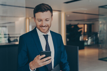 Happy young businessman using smart phone standing in office hallway