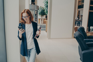 Smiling redhead female business consultant dressed formally holding mobile phone during coffee break