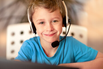 Boy with headphones is sitting in front of a computer monitor. He studies online from home. Portrait of a smiling cute boy in close-up.