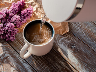 Unrecognizable man preparing coffee. Pouring water into a cup. Morning concept of making tea or coffee on a wooden background with a bouquet of lilacs.