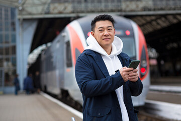Male Asian businessman, at the train station, buys train tickets online, uses a mobile phone and an...