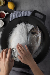 Woman hands covering completely a sea bream with a rock salt layer. The other sea bream is waiting its turn.