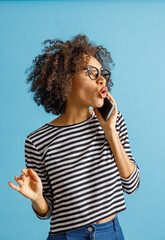 Astonished multiracial lady in striped long sleeve shirt having phone conversation. Isolated on blue background