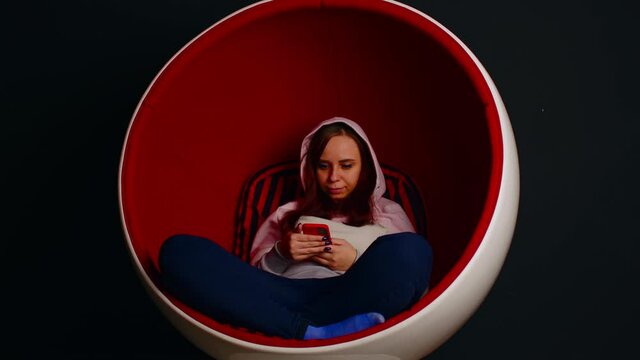 Woman browsing smartphone in egg armchair. Female hipster sitting in white and red egg shaped armchair and messaging on social media via cellphone on black background