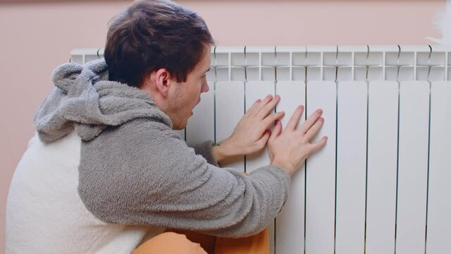 Man warming hands on radiator. Frozen male cringing from cold, warming hands on heating radiator in winter day.