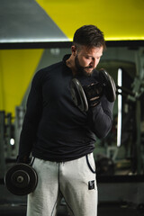 Fototapeta na wymiar Front view portrait of young caucasian man male athlete bodybuilder training at the gym workout using dumbbells biceps curls wearing black shirt dark hair and beard standing weight lifting copy space