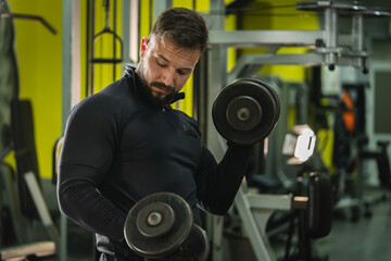 Side view portrait of young caucasian man male athlete bodybuilder training at the gym workout using dumbbells biceps curls wearing black shirt dark hair and beard standing weight lifting copy space
