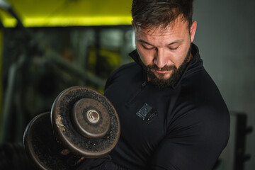 Obraz na płótnie Canvas Side view portrait of young caucasian man male athlete bodybuilder training at the gym workout using dumbbells biceps curls wearing black shirt dark hair and beard standing weight lifting copy space