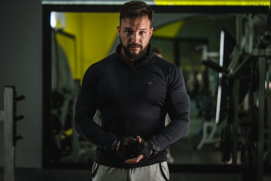 Front View Waist Up Portrait Of Young Adult Caucasian Man With Dark Hair And Beard In Black Gym Suit Looking To The Camera Adjusting Gloves - Confident Modern Male Dramatic Portrait With Copy Space