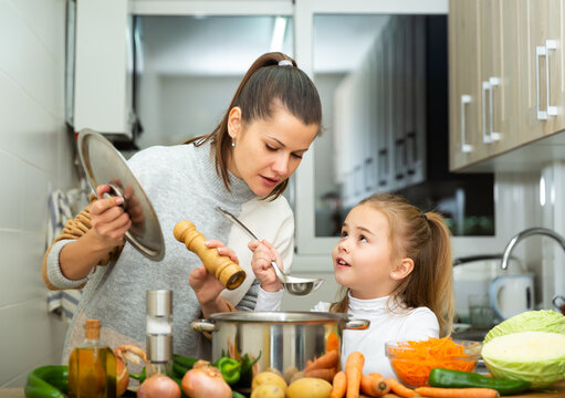 Little daughter helping cooking soup and mother add pepper to pan