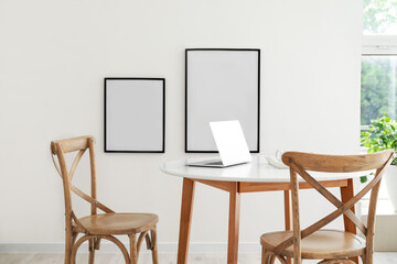 Fototapeta na wymiar Dining table with laptop and blank frames hanging on light wall