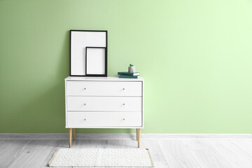 Blank frames and books on chest of drawers near green wall