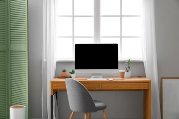 Wooden table with computer, cup of coffee and flowerpots near window in room