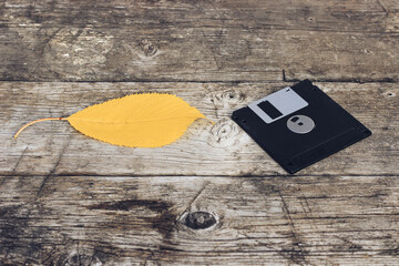 A black floppy disk and a yellow autumn leaf on a wooden table. The concept of preserving autumn impressions