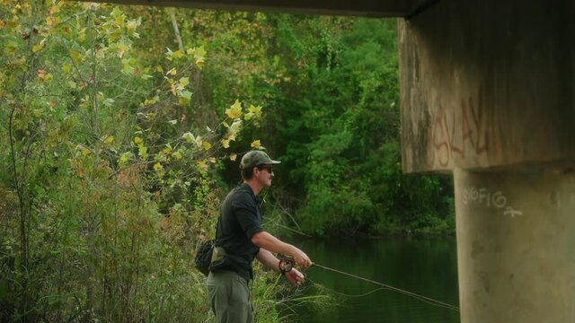 Texas flyfisherman casting flyrod under bridge near a beautiful green river with fall foliage and then stripping rod side profile in slow motion 4k