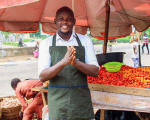 A happy African male trader with an apron standing beside his stall of tomatoes and pepper in a...