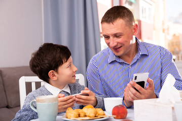 Obraz na płótnie Canvas Cheerful man and his preteen son absorbedly looking at smartphones over cup of tea at home table