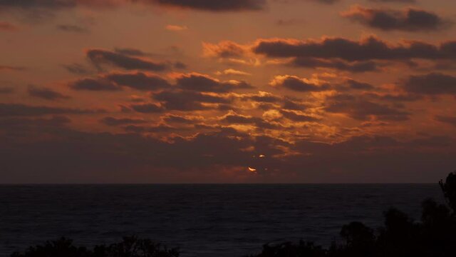 Sunset over Indian Ocean, sun obscured by grey clouds flecked with gold and orange. Time lapse