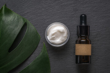 A jar of cream, a large green leaf and a dropper bottle on a black stone background. The concept of natural therapy and cosmetics.
