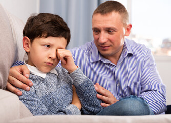 Young father consoling preteen son sitting at sofa after quarrel at home