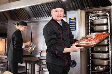 Skilled cook working in professional kitchen, preparing to put pork ribs and sausages into oven