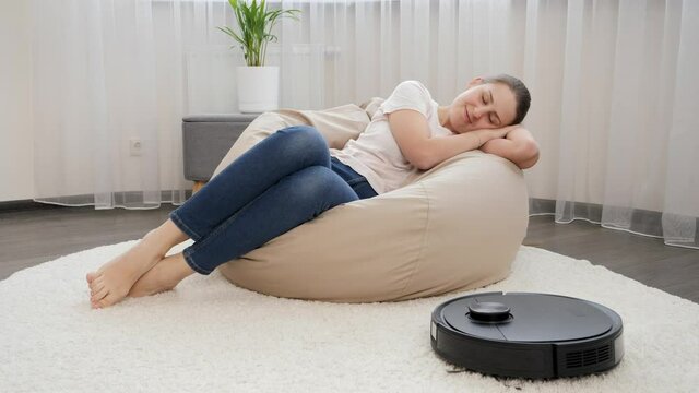 Young housewife relaxing in living room while robot vacuum cleaner doing housework. Concept of hygiene, household gadgets and robots at modern life.