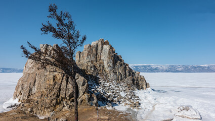 Fototapeta na wymiar A picturesque two-headed rock, devoid of vegetation, rises above a frozen lake. Cracks on steep slopes. There is ice and snow all around. There is a bare tree in the foreground. Clear blue sky. Baikal