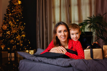 Mother and son are lying on the bed in pajamas with gifts on the background of the Christmas tree.