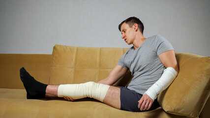 Man with short brunet hair sits on soft sofa in living room feeling frustrated with injured knee in...