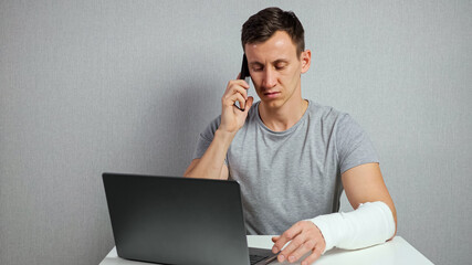 Young brunette-haired man freelancer in grey t-shirt with broken forearm in gypsum band works distantly using laptop and talking via smartphone. - obrazy, fototapety, plakaty