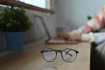 Eyeglasses on wooden table and young woman using laptop in background.