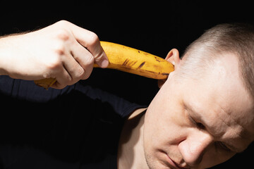 Man holds banana in his ear close-up on black background. Concept of inability to understand others...