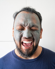 Beauty portrait of an excited matured Asian man standing isolated over white background, wearing a facial cosmetic clay mask.