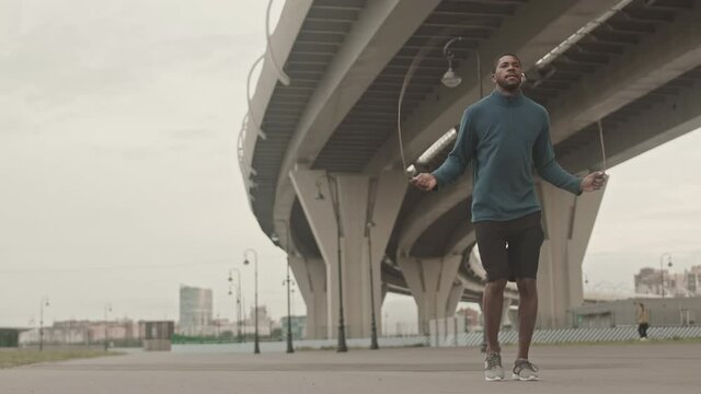 Stab full-length shot with slowmo of young Black man in activewear skipping rope during outdoor training on cold morning