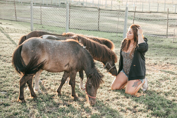 Young woman is sitting next to three grazing ponies . Life on farm. Agrotourism. Beautiful animals...