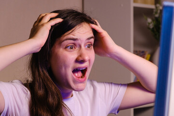 A woman screams and grabs her head with her hands while looking at a TV or computer screen.