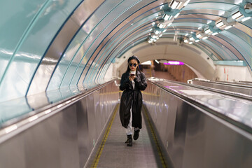Stylish asian girl blogger in leather trench and eyeglasses on escalator using smartphone reading followers comments and posting photos to social media. Chinese woman influencer communicate online