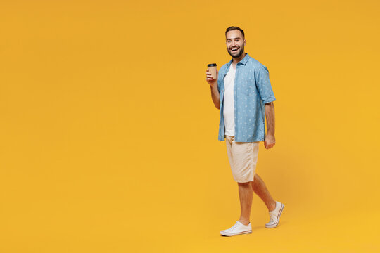 Full Body Young Happy Man In Blue Shirt White T-shirt Hold Takeaway Delivery Craft Paper Brown Cup Coffee To Go Walking Isolated On Plain Yellow Background Studio Portrait. People Lifestyle Concept.